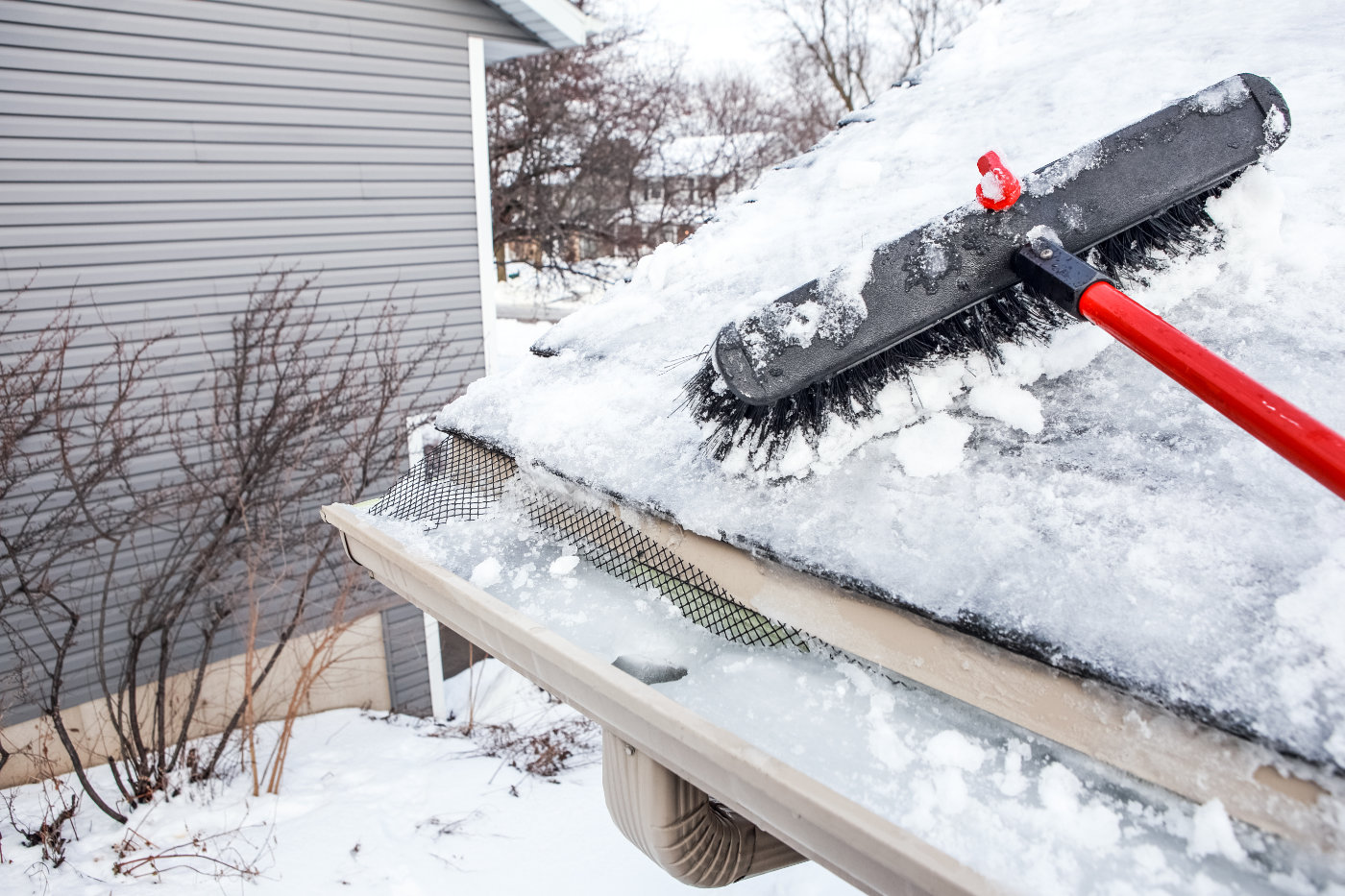 Broom raking snow off of roof, with ice dam build-up in gutter and eavestroughs.