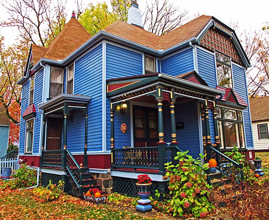 A charming blue Victorian house with a large wraparound porch, ornate trim, and colorful fall foliage in the yard.