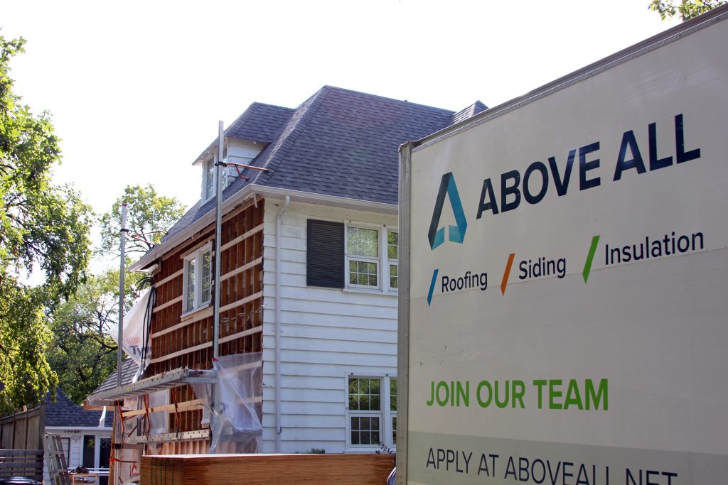 A white truck with the logo and contact info for Above All, a roofing, siding, and insulation company, is parked in front of a house under renovation.