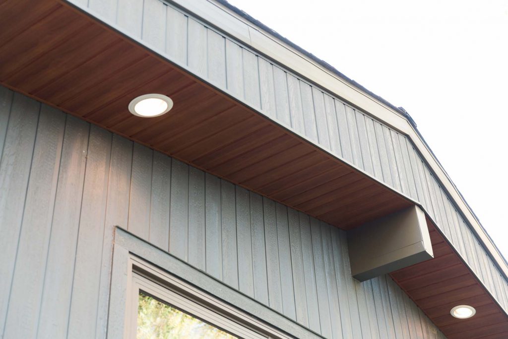 A modern home exterior featuring dark grey vertical siding, wood accents, and recessed lighting under the soffit.