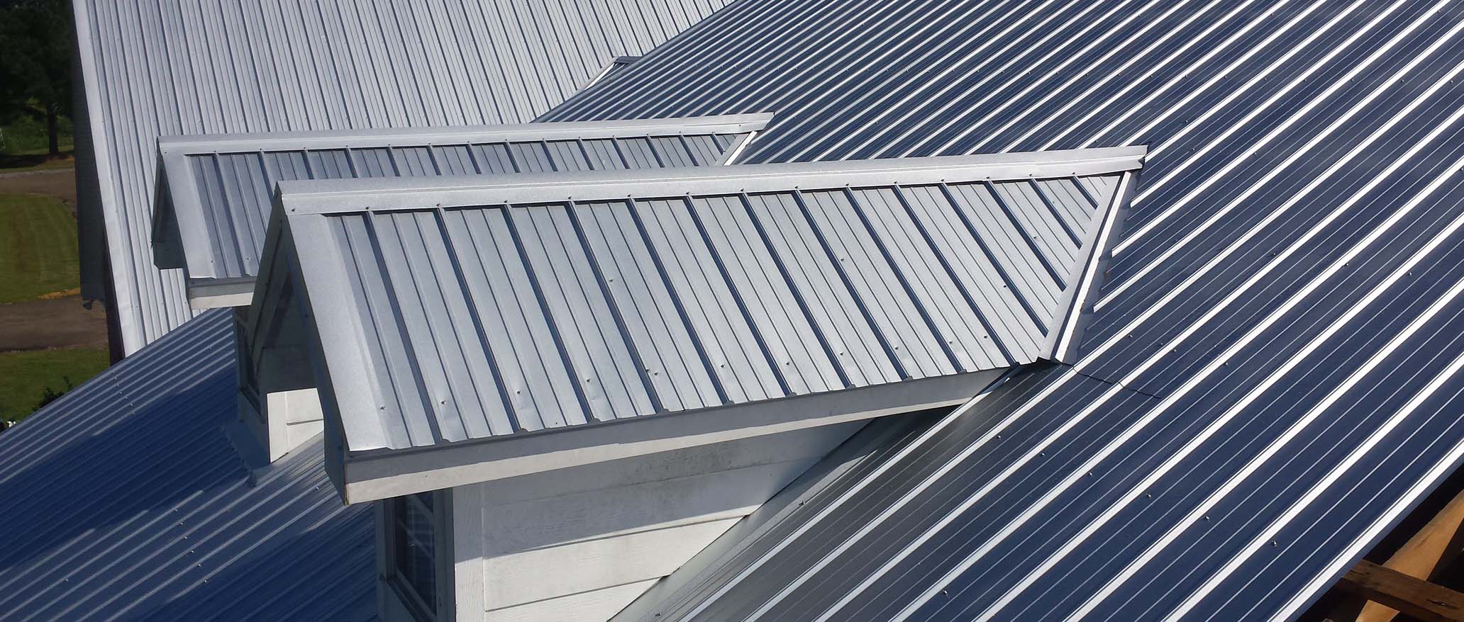 A light grey, ribbed metal roof with two dormers on a sunny day.