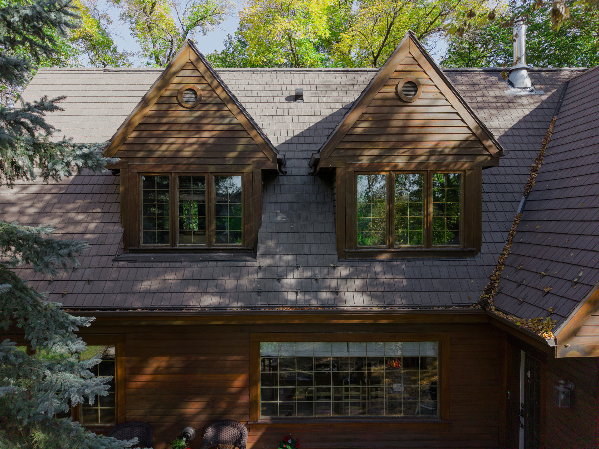 Wood home with twin dormers and shingle roof surrounded by trees and shade.