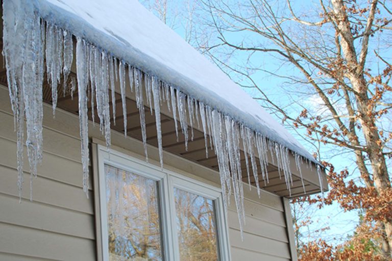 Icicles hanging from the edge of a snow-covered roof, showing ice buildup along the eaves of a house in winter.